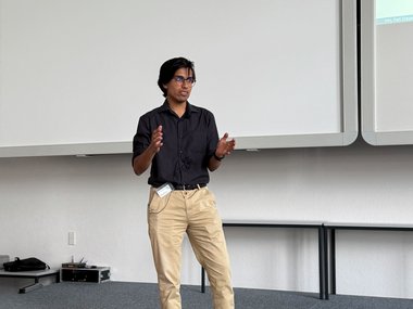 Matter to Life PhD candidate Pratulya in a dark shirt and khaki pants gestures on a stage, with a large projection screen behind them.
