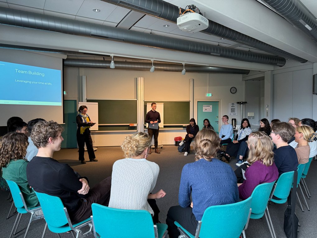 Seminar room full of people engaged in conversation, two speakers in front of a presentation on team building displayed on a screen.