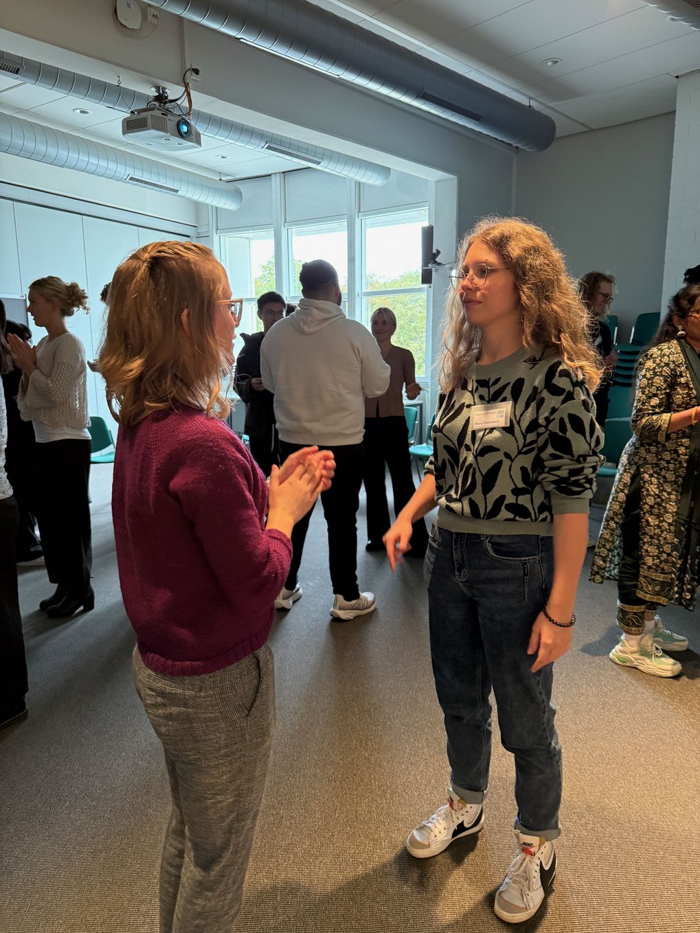 People engaged in conversation in a bright room with chairs stacked in the corner.