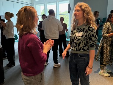 People engaged in conversation in a bright room with chairs stacked in the corner.