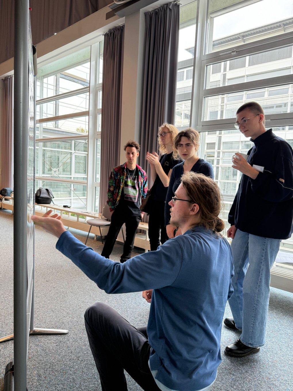 Individuals gathered in a spacious room with floor-to-ceiling windows, engaging in a discussion near a presentation board.