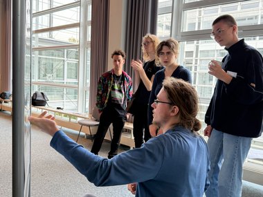 Individuals gathered in a spacious room with floor-to-ceiling windows, engaging in a discussion near a presentation board.