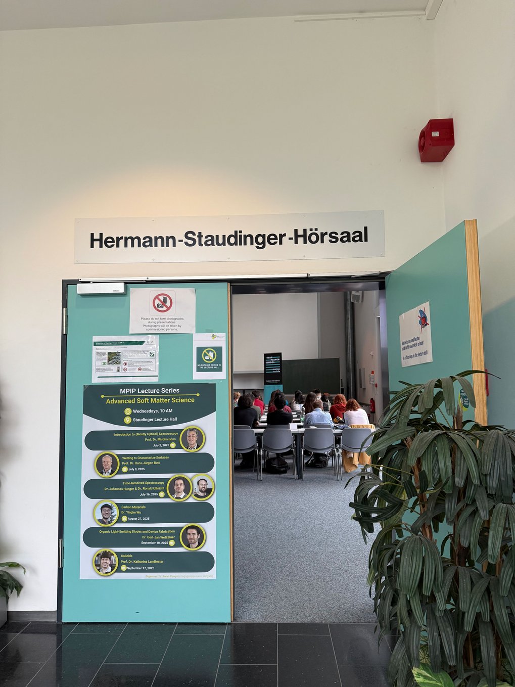 Open door to Hermann-Staudinger Lecture Hall displaying lecture series poster; audience seated inside the hall.