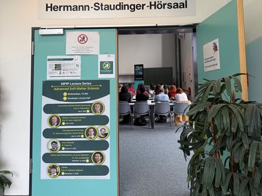 Open door to Hermann-Staudinger Lecture Hall displaying lecture series poster; audience seated inside the hall.