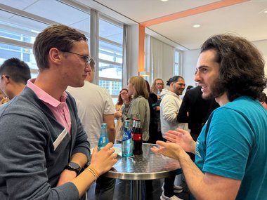 Individuals converse at a high table, surrounded by beverages in a well-lit space.
