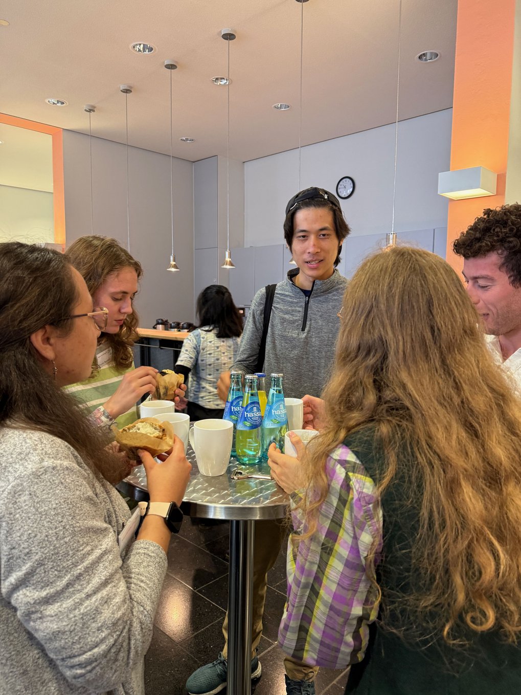 Several people gather around a tall table with bottled water, mugs, and sandwiches in a brightly lit room.