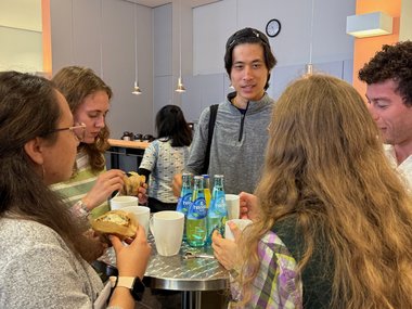 Several people gather around a tall table with bottled water, mugs, and sandwiches in a brightly lit room.