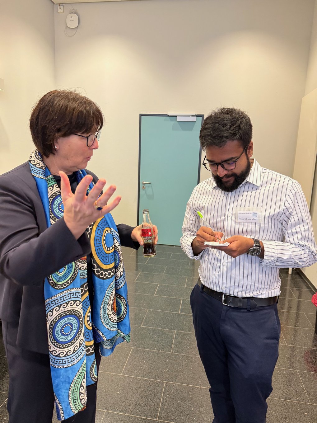 A woman with a vibrant blue scarf holds a bottle while conversing with a man jotting down notes on a notepad.