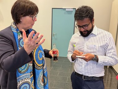 A woman with a vibrant blue scarf holds a bottle while conversing with a man jotting down notes on a notepad.