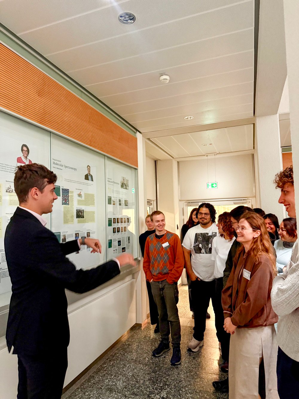 A presenter engages an audience in a corridor with educational panels and a ceiling with recessed lighting.