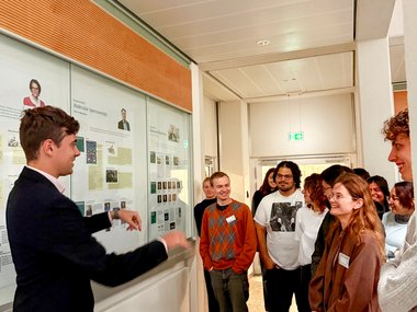 A presenter engages an audience in a corridor with educational panels and a ceiling with recessed lighting.