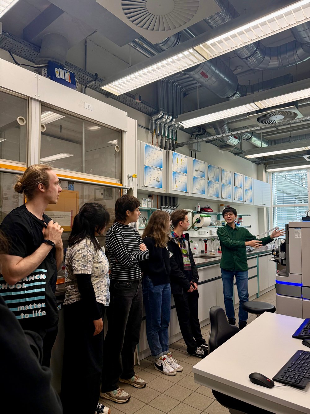 In a modern laboratory, a group attentively listens to a presenter who is explaining the function of a complex scientific instrument, surrounded by technical equipment and detailed diagrams on the walls.In a modern laboratory, a group attentively listens to a presenter who is explaining the function of a complex scientific instrument, surrounded by technical equipment and detailed diagrams on the walls.