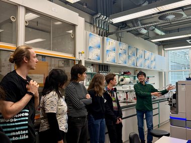 In a modern laboratory, a group attentively listens to a presenter who is explaining the function of a complex scientific instrument, surrounded by technical equipment and detailed diagrams on the walls.In a modern laboratory, a group attentively listens to a presenter who is explaining the function of a complex scientific instrument, surrounded by technical equipment and detailed diagrams on the walls.