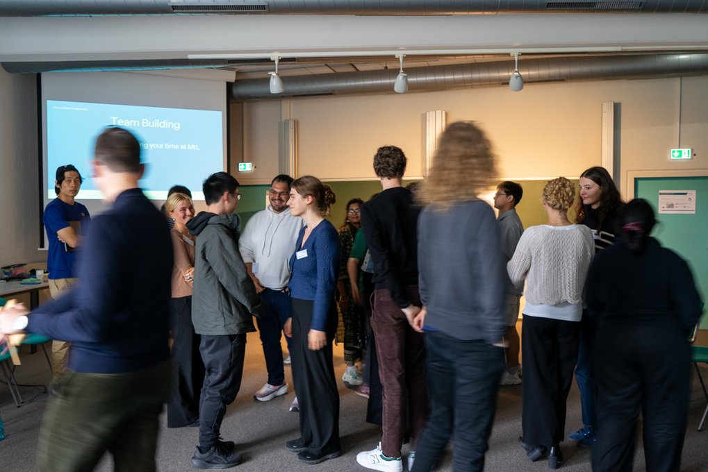 Participants gather in a room, focusing on a team-building exercise. A screen displays related information. Some converse in small groups.