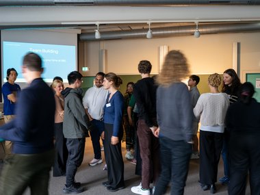 Participants gather in a room, focusing on a team-building exercise. A screen displays related information. Some converse in small groups.