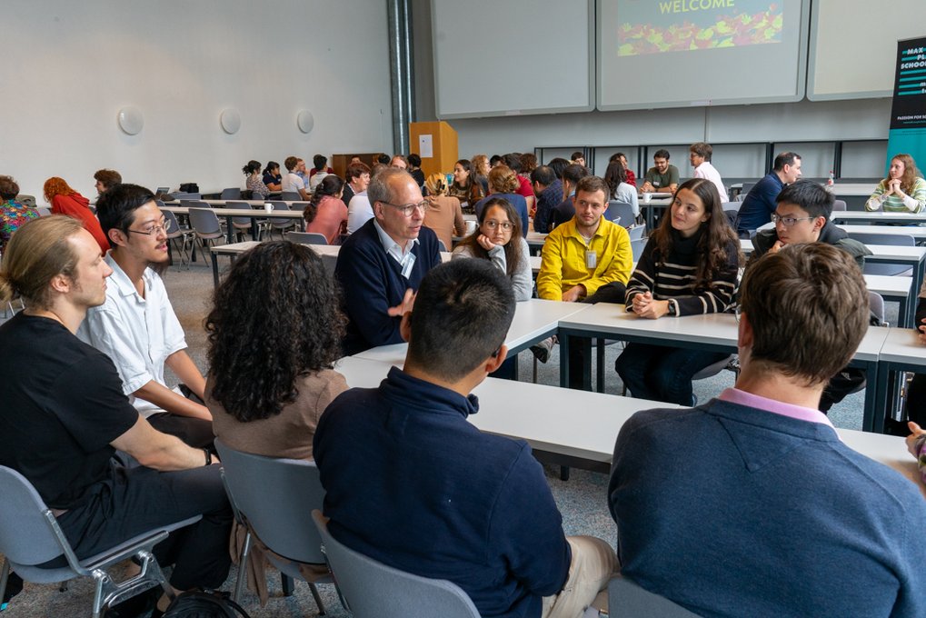 Several groups of people are seated at rectangular tables in a conference room, engaging in discussions, with a presentation screen in the background.