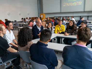 Several groups of people are seated at rectangular tables in a conference room, engaging in discussions, with a presentation screen in the background.