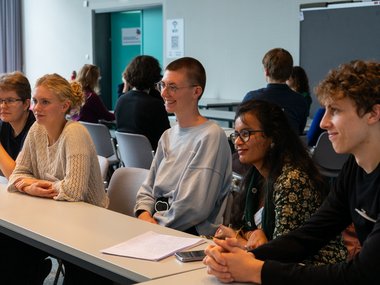 In a modern conference room, several individuals are seated at tables, some with notebooks and mobile devices, while a presentation takes place.