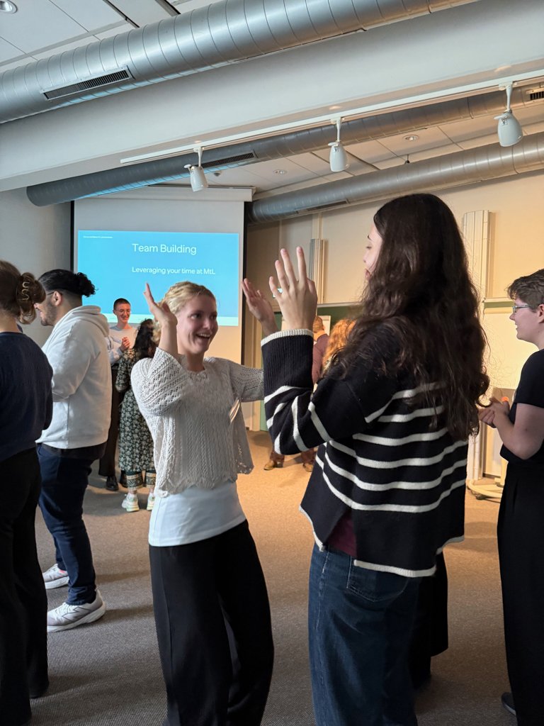 New PhD candidates participating in a lively team-building exercise in a seminar room, raising their hands toward each other while others engage in activities in the background. A presentation slide titled “Team Building” is visible on the screen.