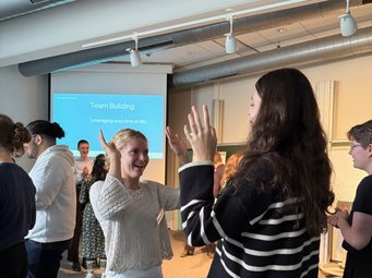 High spirits and teamwork: our new PhD candidates during a group-building workshop by Mighty Real. New PhD candidates participating in a lively team-building exercise in a seminar room, raising their hands toward each other while others engage in activities in the background. A presentation slide titled “Team Building” is visible on the screen.