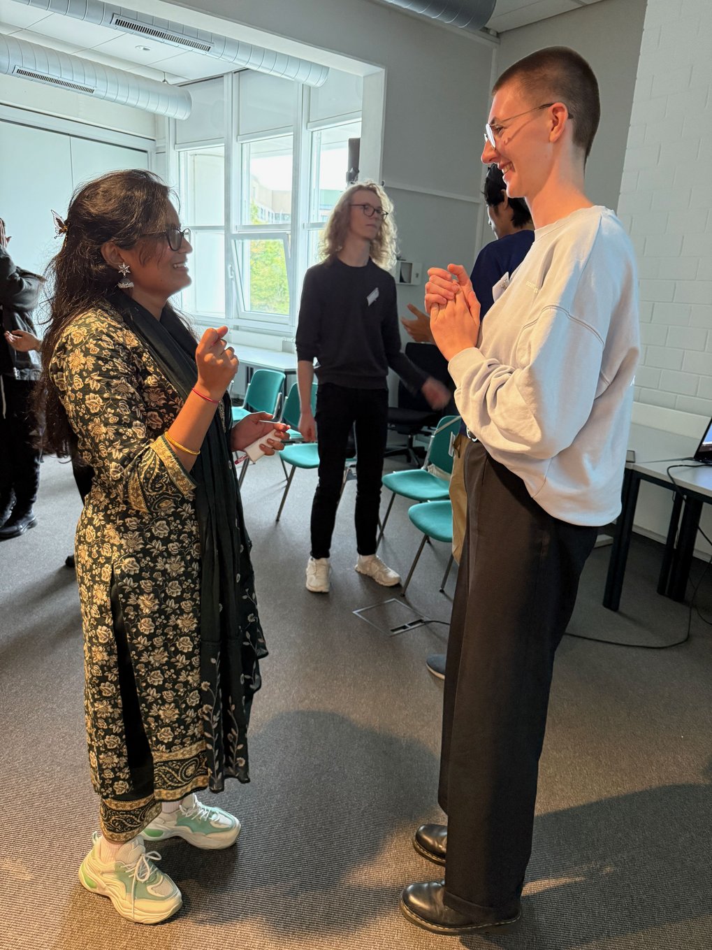Two new PhD candidates talking and laughing during a team-building exercise in a bright seminar room, with other participants visible in the background.