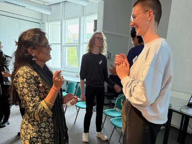 Two new PhD candidates talking and laughing during a team-building exercise in a bright seminar room, with other participants visible in the background.