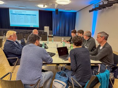 A group of people sits at a conference table, utilizing laptops and discussing ideas. The room features a presentation screen displaying session details and several empty chairs.