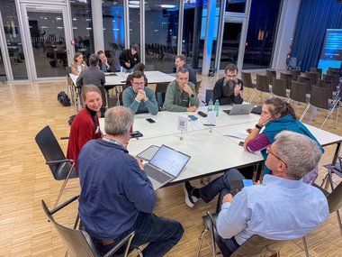 Several people gathered around tables in a modern meeting room. Some are using laptops, while others have mobile phones. Bottles of water are on the tables.