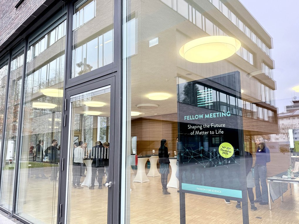 View through glass windows reveals attendees at a science meeting inside a modern building.