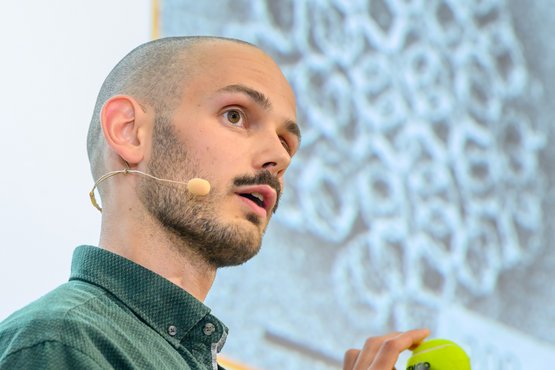 Christoph Karfusehr with a microphone, wearing a green shirt, holds a tennis ball while standing by a screen displaying an microscope image.