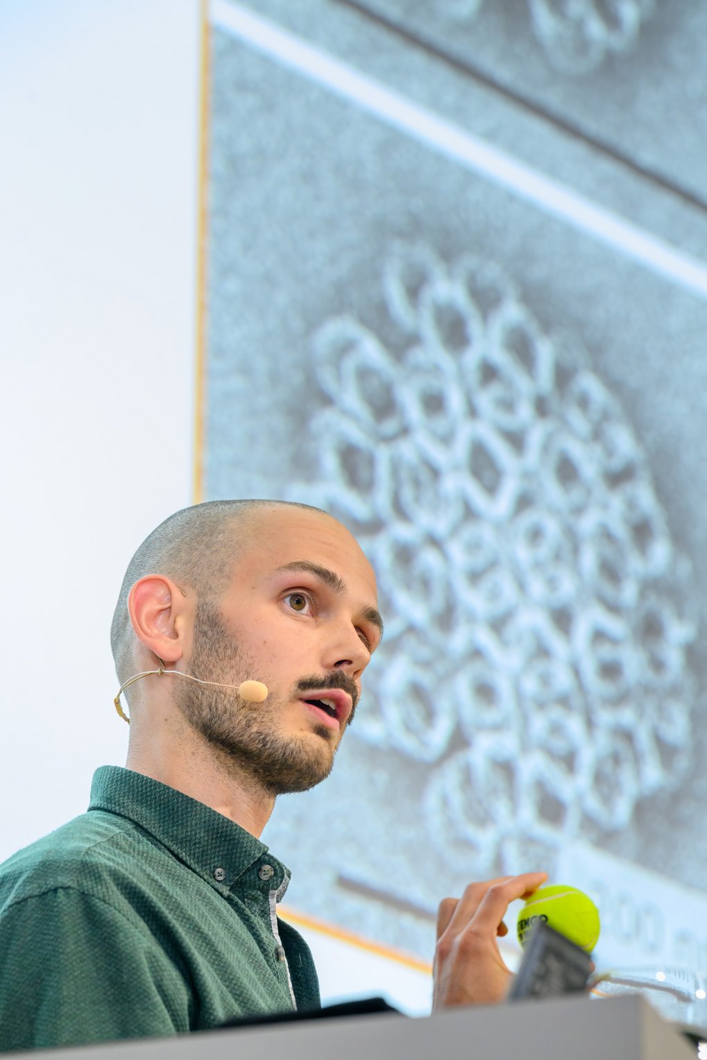 Christoph Karfusehr with a microphone, wearing a green shirt, holds a tennis ball while standing by a screen displaying an microscope image.