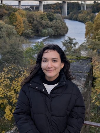 Laura stands amidst a picturesque autumn scene featuring a river winding through vibrant foliage, with a large bridge spanning across in the background.