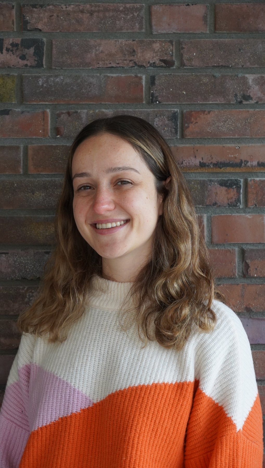 Aysecan Ünal wearing a sweater with orange and purple stripes, standing against a brick wall.