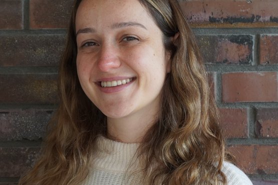 Aysecan Ünal, die einen Wollpullover mit orangefarbenen, weißen und rosa Streifen trägt und vor einer strukturierten Backsteinwand steht.