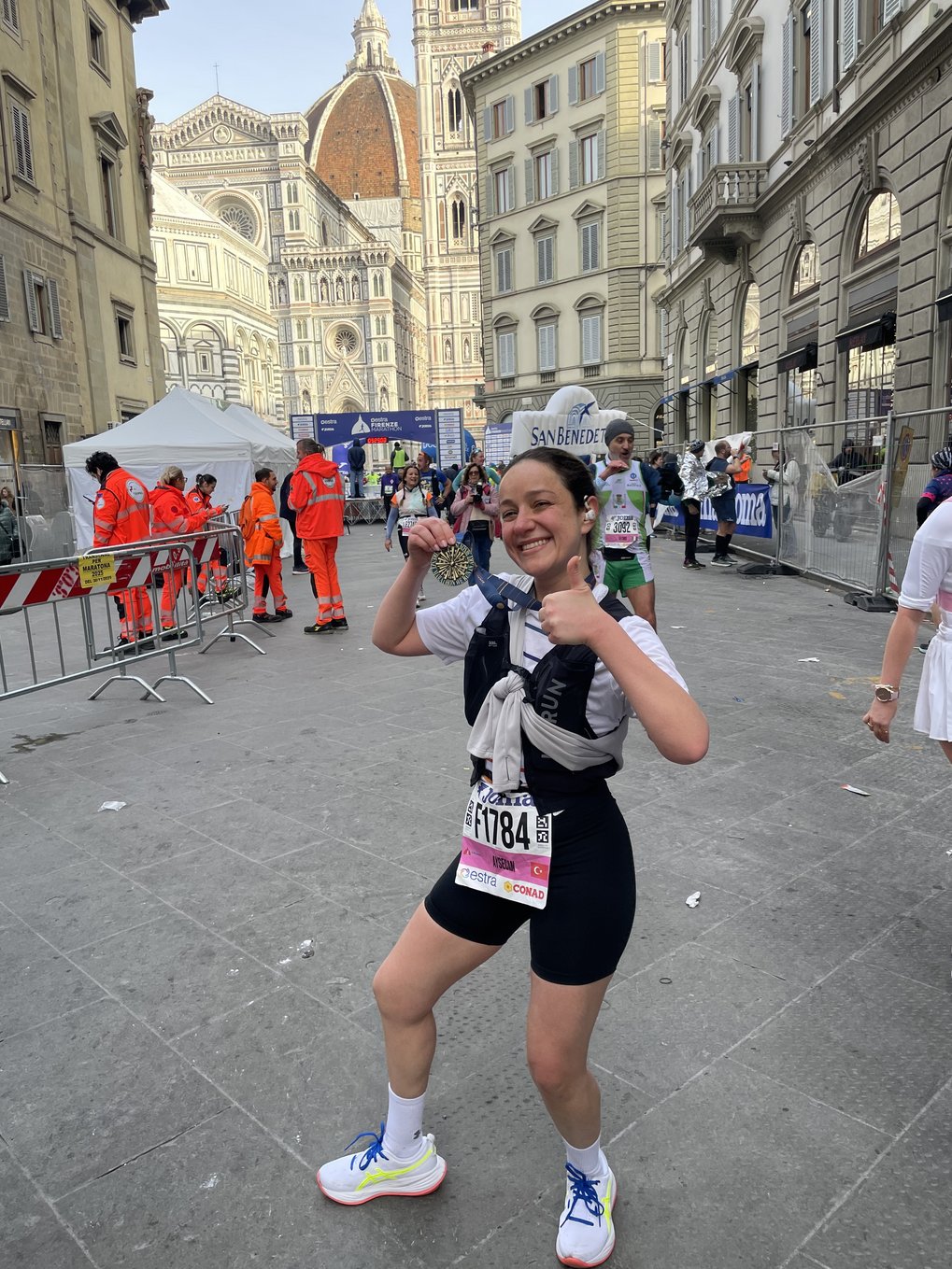 Aysecan, dressed in sportswear, proudly displays a medal in front of Florence Cathedral. Other runners, spectators, and emergency personnel can be seen in the background.