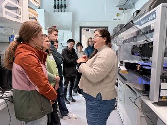 Candice Jones, head of the MaxGenesys biofoundry showed us equipment, and explained the workflows to automate synthetic biology research Several individuals in a laboratory, focusing on a large piece of scientific equipment, with one person explaining something.