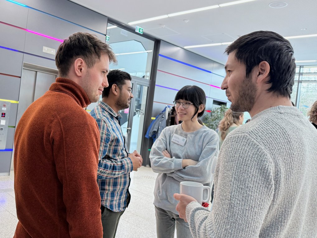 Four people are gathered in a contemporary space with colorful lighting, holding a discussion. One person holds a white mug.