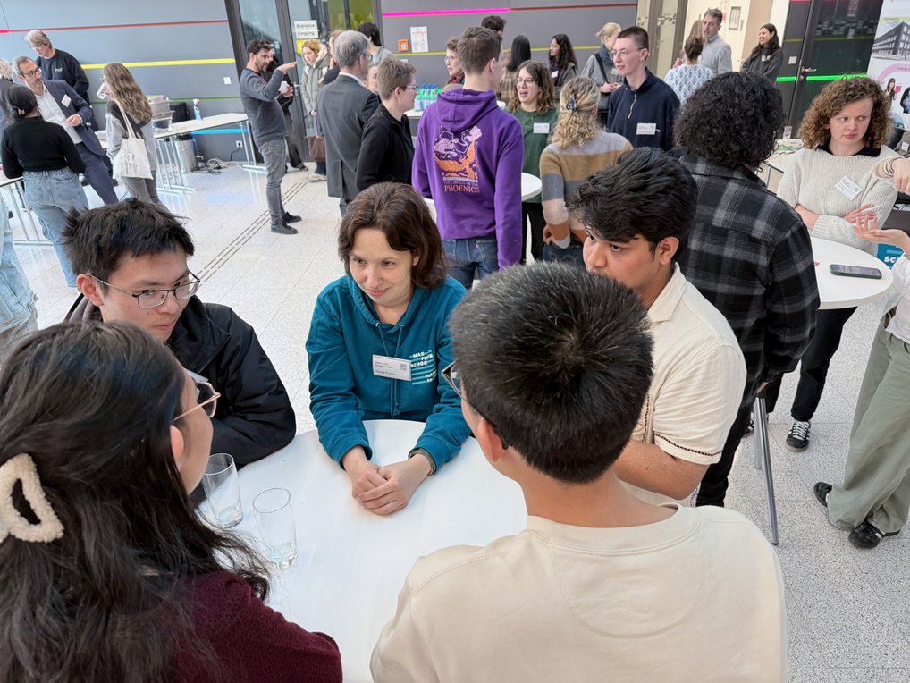 Symposium participants during a coffee break in a modern interior: People are chatting at tables; some are wearing name tags, while others are holding drinks.

