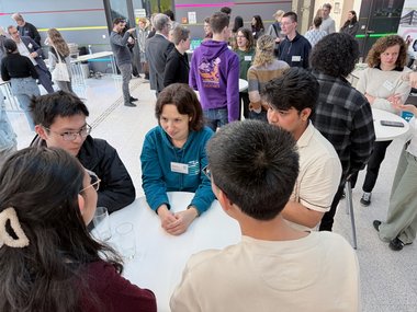 Symposium participants during a coffee break in a modern interior: People are chatting at tables; some are wearing name tags, while others are holding drinks.