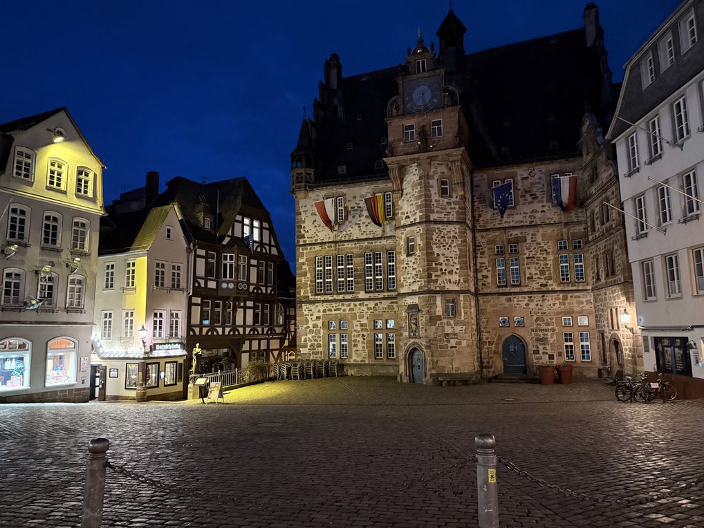The illuminated historic Marburg City Hall on a cobblestone square with flags at dusk.