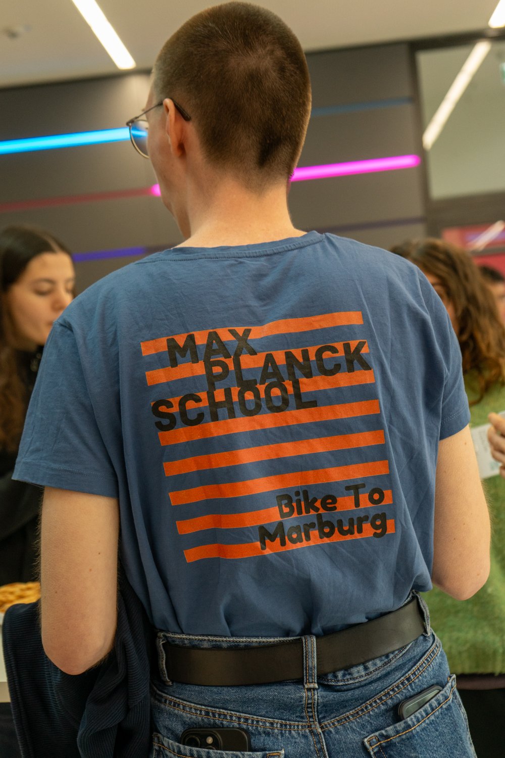 A person stands in a contemporary room, wearing a blue shirt with orange stripes featuring "Max Planck School" and "Bike to Marburg" text.