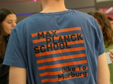 A person stands in a contemporary room, wearing a blue shirt with orange stripes featuring "Max Planck School" and "Bike to Marburg" text.