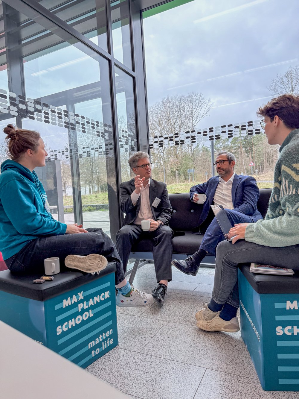 Four people in a casual discussion in a modern room with large windows.