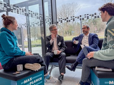Four people in a casual discussion in a modern room with large windows.