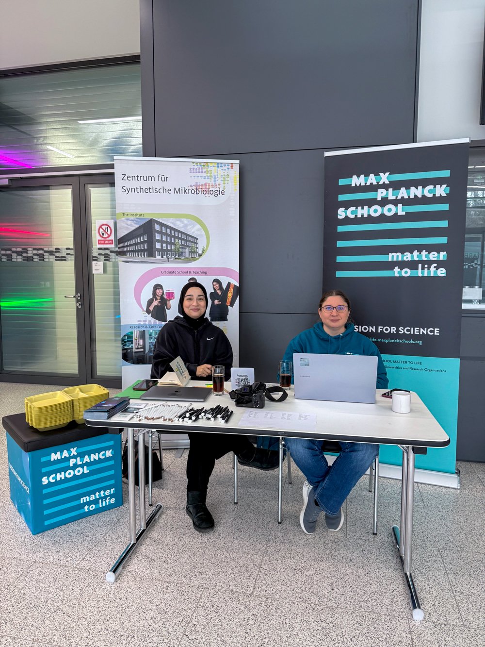 A reception desk decorated with banners from the Max Planck School Matter to Life and the Center for Synthetic Microbiology, where two Matter to Life program coordinators are seated. On the desk are a laptop, a camera, pens, and brochures.