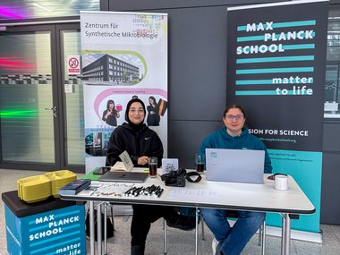 A reception desk decorated with banners from the Max Planck School Matter to Life and the Center for Synthetic Microbiology, where two Matter to Life program coordinators are seated. On the desk are a laptop, a camera, pens, and brochures.