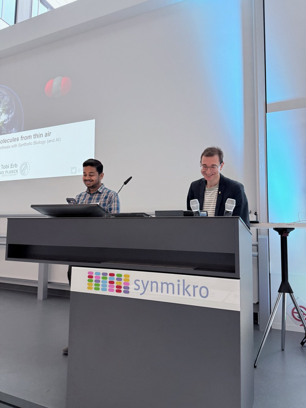 Two individuals are at a podium labeled synmikro, preparing for a presentation. Behind them, a screen displays a talk on synthesizing molecules using synthetic biology and AI.