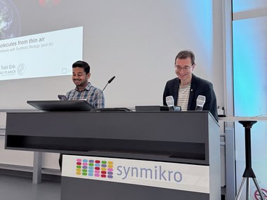 Two individuals are at a podium labeled synmikro, preparing for a presentation. Behind them, a screen displays a talk on synthesizing molecules using synthetic biology and AI.