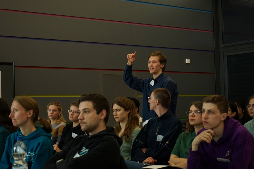 In a lecture hall, several people are seated, while one individual stands and gestures with a raised hand, possibly asking a question.