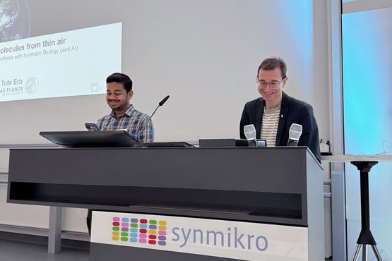 Two individuals are at a podium labeled synmikro, preparing for a presentation. Behind them, a screen displays a talk on synthesizing molecules using synthetic biology and AI.